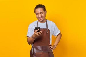 Portrait of Asian young man wearing apron smiling broadly looking at incoming message on smartphone on yellow background photo