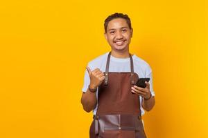 Cheerful young Asian man holding mobile phone and pointing fingers aside at copy space isolated over yellow background photo