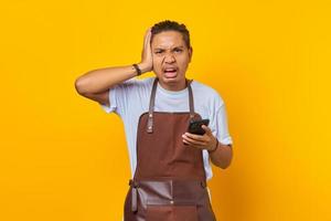 Portrait of shocked young Asian man hand on head with disappointed expression and holding smartphone on yellow background photo