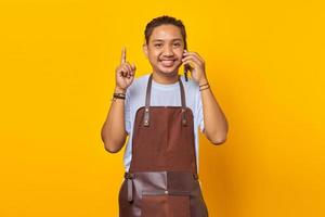 Portrait of smiling Asian young man talking on smartphone and finger pointing up. yellow background photo