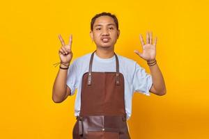 Portrait of confident handsome Asian young man wearing apron pointing with finger number seven with angry expression photo