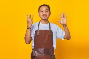 Portrait of confident handsome Asian young man wearing apron pointing with number eight finger with angry expression photo