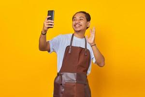 Portrait of cheerful asian young man wearing apron making video call on smartphone isolated on yellow background photo