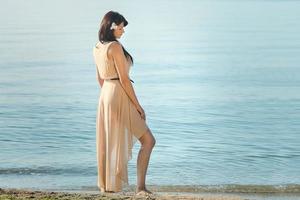 girl with black hair in a beige dress on the shore of the ocean photo