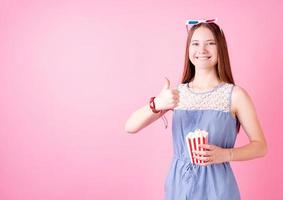 Smiling teenager girl wearing 3D glasses eating popcorn isolated on pink background showing thumb up photo