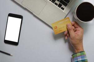 Vista superior de un hombre que sostiene un teléfono móvil con tarjeta de crédito simulacro de pantalla en blanco en blanco y tecnología de comunicación empresarial de computadora y café, concepto de compras en línea.