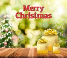 Feliz Navidad palabra brillo rojo y regalo dorado sobre mesa de madera marrón con árbol de Navidad bokeh borrosa foto