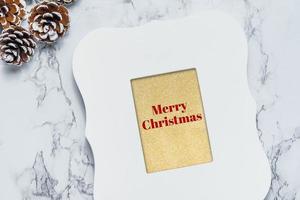 Merry christmas text in vintage white picture frame with pine cones and ribbon on white marble table photo