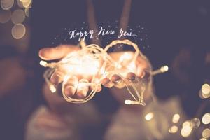 Feliz año nuevo palabra resplandeciente sobre mano con cadena de luz de fiesta bokeh en filtro vintage foto