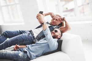 Two stylish young friends laying on the sofa and taking selfie. Blonde guy showing biceps