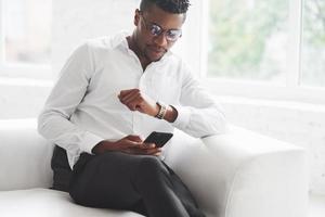 New deal is coming. Checking the time. Young afro american guy in office wear sitting on the white sofa photo