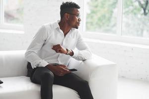 mirando hacia un lado. Joven afroamericano en ropa de oficina sentado en el sofá blanco