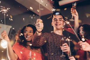 gozo y felicidad. amigos multirraciales celebran el año nuevo y sostienen luces de bengala y vasos con bebida