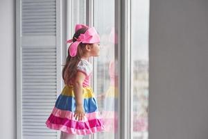 Childish curiosity. Photo of young cute girl in colorful wear standing near the window and looking outside