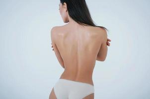 Hugging herself. Girl with perfect body in white underwear turning her back to the white background in the studio photo