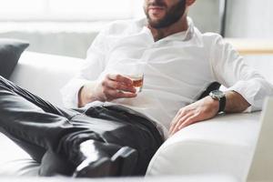tener un merecido descanso. Foto recortada del joven empresario en ropa clásica sentado en el sofá con vaso de whisky en la mano