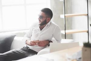 Photo from the side. Photo of young bearded businessman in sunglasses and whiskey in the hand sit on the white sofa