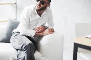 Foto de joven empresario barbudo en gafas de sol y whisky en la mano sentarse en el sofá blanco