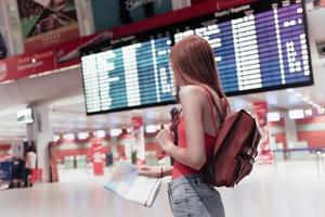 Mujer joven en el aeropuerto con mapa en manos y tablero de información en el fondo está mirando hacia el lado