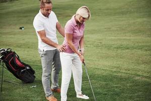 Girl playing golf and hitting by putter on green. Her teacher helps to explore the technique and make her first strikes photo