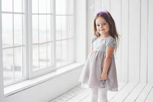 linda niña en vestido de pie en la habitación blanca cerca de las ventanas