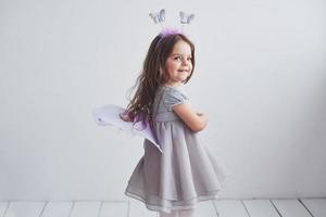 humor alegre y sonrisa sincera. Encantadora niña en traje de hadas de pie en la habitación con fondo blanco.