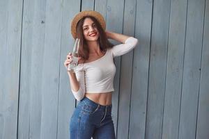 mujer positiva con estilo. Retrato de una hermosa joven con sombrero en la cabeza y una botella en la mano de pie contra la pared de madera foto