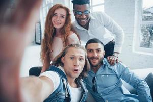 camarógrafos preguntándose. Alegres jóvenes amigos tomando autorretratos en el sofá y el interior blanco foto