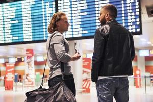 discusión sobre la planificación de un viaje. Foto de dos camaradas que se encuentran en el aeropuerto cerca del sistema de visualización de información de vuelo