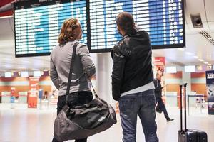 Man shows time of plane arrival. Photo of two comrades situating in airport near flight information display system