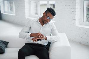 admira el nuevo reloj de pulsera. Elegante chico afroamericano con ropa clásica y gafas está sentado en la cama foto