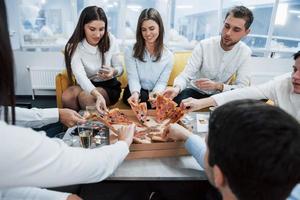 tomando las rodajas. comiendo pizza. celebrando un trato exitoso. jóvenes trabajadores de oficina sentados cerca de la mesa con alcohol foto