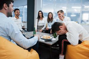 Guy laughing about his loss with hands on the face. Celebrating successful deal. Young office workers sitting near the table with alcohol photo