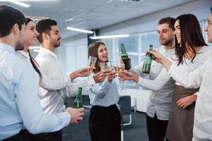 Sincere smiles. Standing and knocking the bottles and glass. In the office. Young people celebrate their success photo