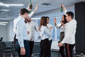 Making a toast. Photo of young team in classical clothes celebrating success while holding drinks in the modern good lighted office