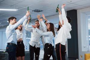 finalmente lo conseguimos. Foto de equipo joven en ropa clásica celebrando el éxito mientras sostiene bebidas en la moderna oficina bien iluminada