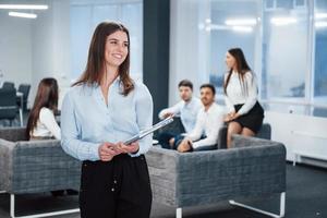 Smiling and looking to the side. Portrait of young girl stands in the office with employees at background photo