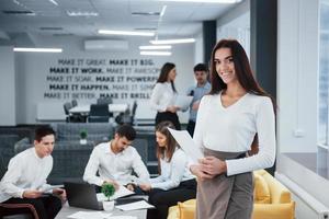 Beautiful brunette. Portrait of young girl stands in the office with employees at background photo