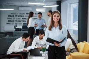 sonriendo mientras sostiene el bloc de notas. Retrato de joven se encuentra en la oficina con empleados en segundo plano. foto