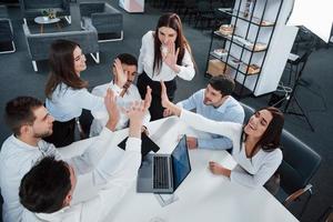 Giving high five to each other. Top view of office workers in classic wear sitting near the table using laptop and documents photo