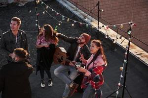 Look, there's a camera up there. Top view of a young friends that drinking, playing guitar and have fun at the roof with decorative colorful light bulbs photo