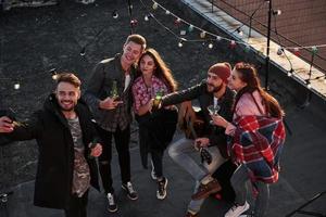 captura el momento. Vista superior de unos jóvenes amigos que se toman selfie mientras se divierten en el techo con bombillas decorativas de colores foto