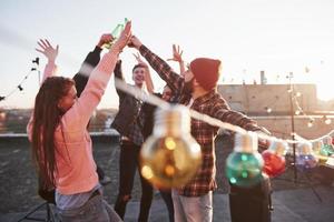 Blurred light bulbs in the front of camera. Holidays on the rooftop. Cheerful group of friends raised their hands up with alcohol photo