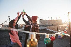 Beautiful sunshine. Holidays on the rooftop. Cheerful group of friends raised their hands up with alcohol photo
