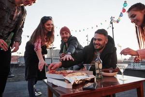 Guy in the red hat makes crazy hungry face and tries to reach that fresh product. Eating pizza at the rooftop party. Good friends have weekend with some delicious food and alcohol photo