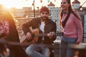 concentrarse en jugar. El guitarrista se sienta y canta para sus amigos en la azotea con bombillas decorativas de colores. foto