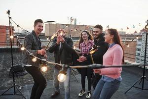 Guy in the red hat imagines that he is a dragon by tying to make flame bigger. Playing with sparklers on the rooftop. Group of young beautiful friends photo