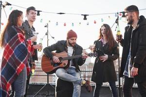 Sing your favorite song. Five young friends have party with beer and guitar at the rooftop photo