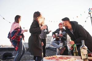 Someone said good joke. Cheerful young people smiling and drinking at the rooftop. Pizza and alcohol on the table. Guitar player