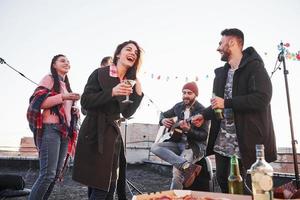 Guy sing funny song. Cheerful young people smiling and drinking at the rooftop. Pizza and alcohol on the table. Guitar player photo